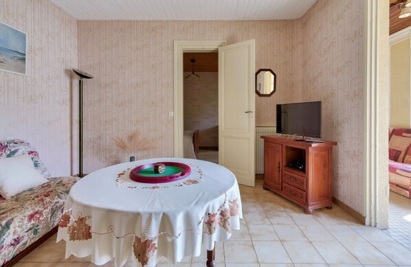 A room with a bed, a table with a decorative tablecloth, a television on a wooden cabinet, and a door leading to another room.