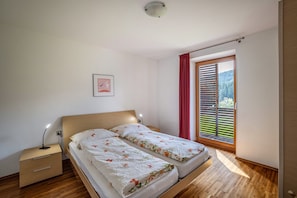 A bedroom with a bed, a nightstand, a bedside lamp, a window with blinds, and a view of greenery outside.