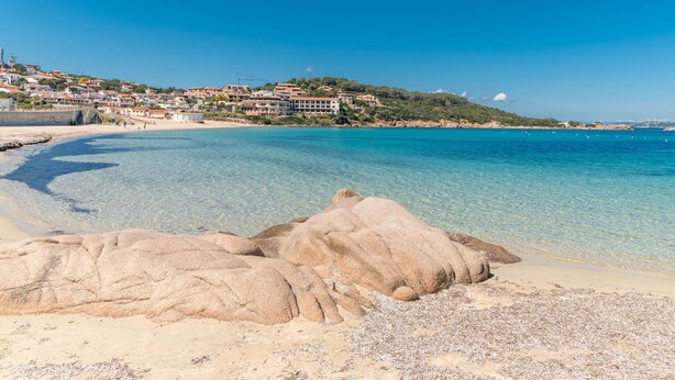 Une ville côtière aux eaux d’un bleu limpide, avec une plage de sable et de gros rochers au premier plan.