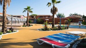 A pool area with lounge chairs, palm trees, and a building with a red roof. - Mobile Home 'Mobil-home Camping Mar Estang' with Lake View, Shared Pool & Air Conditioning (Canet-en-Roussillon)