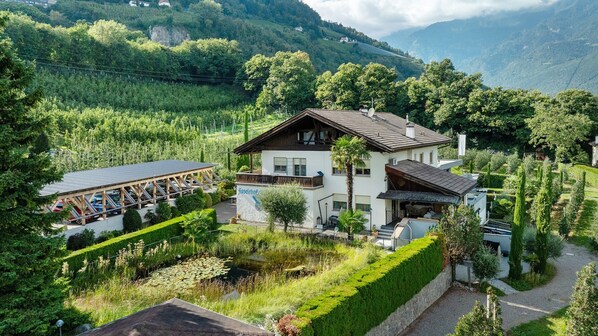 Un chalet de dos plantas con balcón, rodeado de abundante vegetación y un estanque.