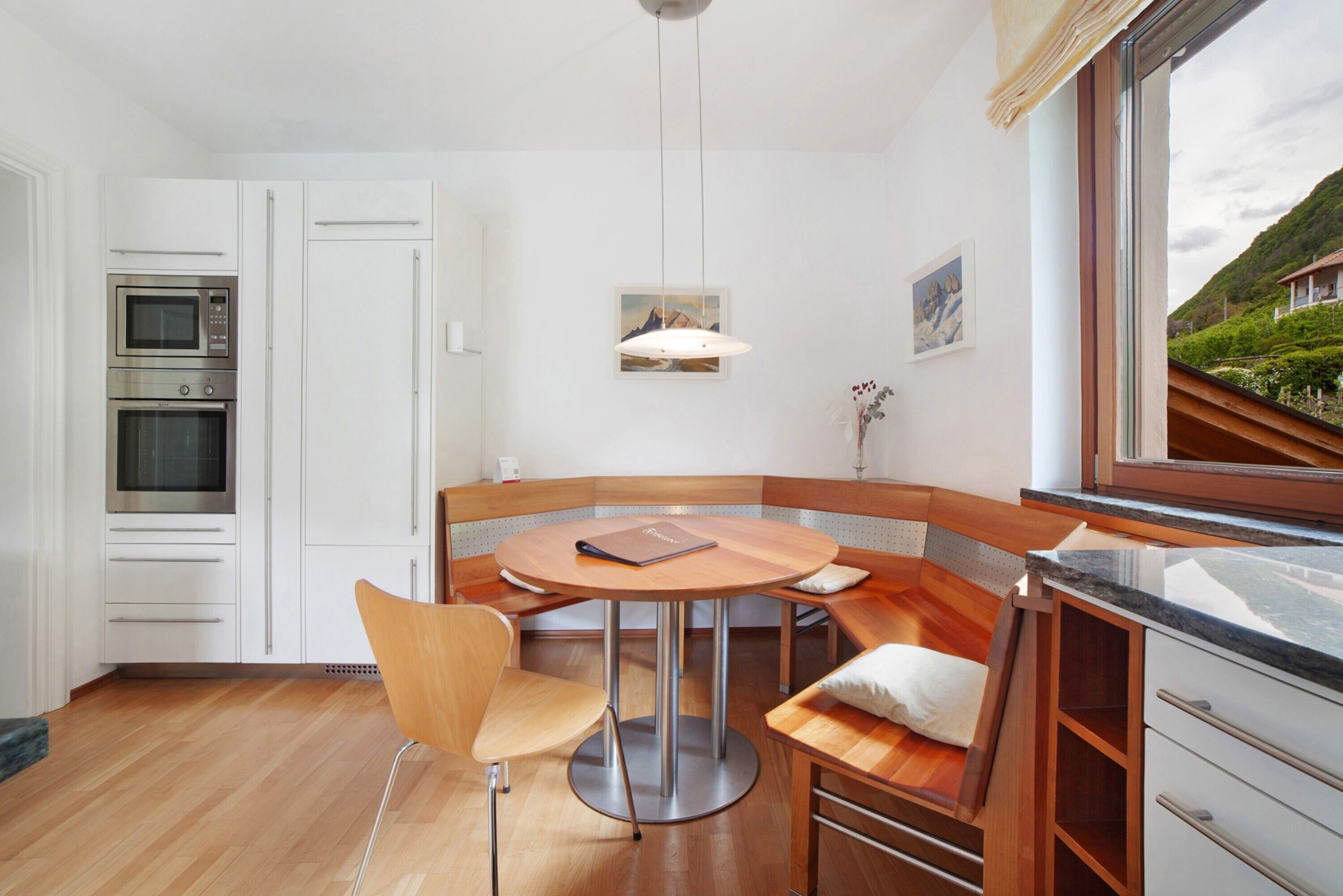 Eine moderne Küche mit eingebauten Geräten, ein Essbereich mit rundem Tisch und Sitzbank sowie ein Blick auf eine gebirgige Landschaft durch ein Fenster.