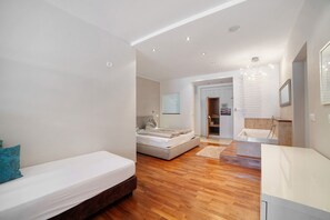 Une chambre moderne comprenant un lit, un petit coin salon, une salle de bain avec baignoire et un couloir menant à une autre pièce.