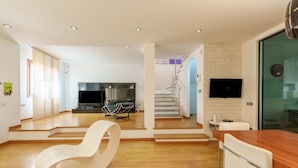 A modern living room with a white rocking chair, a wooden dining table, and a staircase leading to an upper level. - Dimora Smeralda Golfo Aranci – Central Location and Beach with Private Terrace (Golfo Aranci)