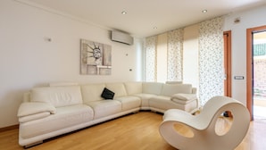 A modern living room with a sectional sofa, a unique white chair, and a wall-mounted air conditioner. - Dimora Smeralda Golfo Aranci – Central Location and Beach with Private Terrace (Golfo Aranci)