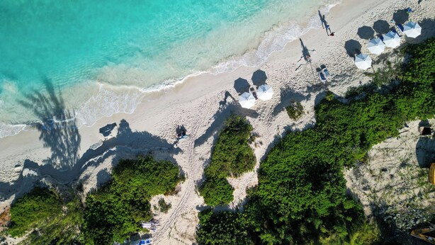 Vue aérienne d’une plage avec une eau turquoise et limpide, un rivage sablonneux, ainsi que plusieurs parasols et chaises longues.