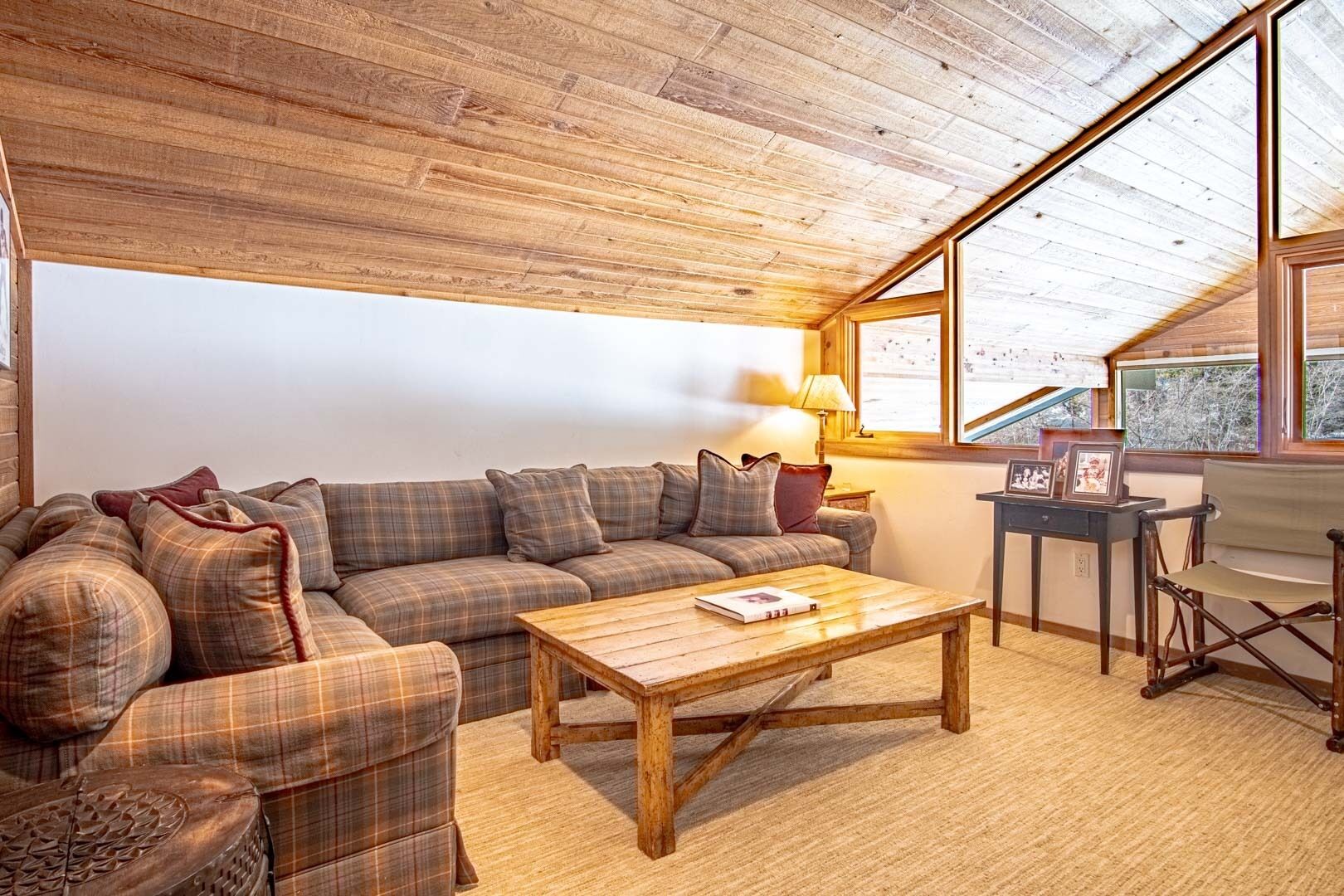 Ein gemütliches Wohnzimmer mit Holzbalkendecke, einem Karomuster-Sofa, einem Holztisch und einem Beistelltisch mit Lampe und Bilderrahmen.