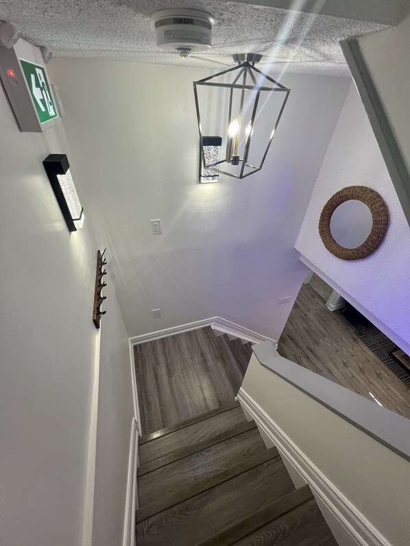 A staircase with wooden steps and a white wall. There is a light fixture above the stairs and a wall-mounted light on the left.