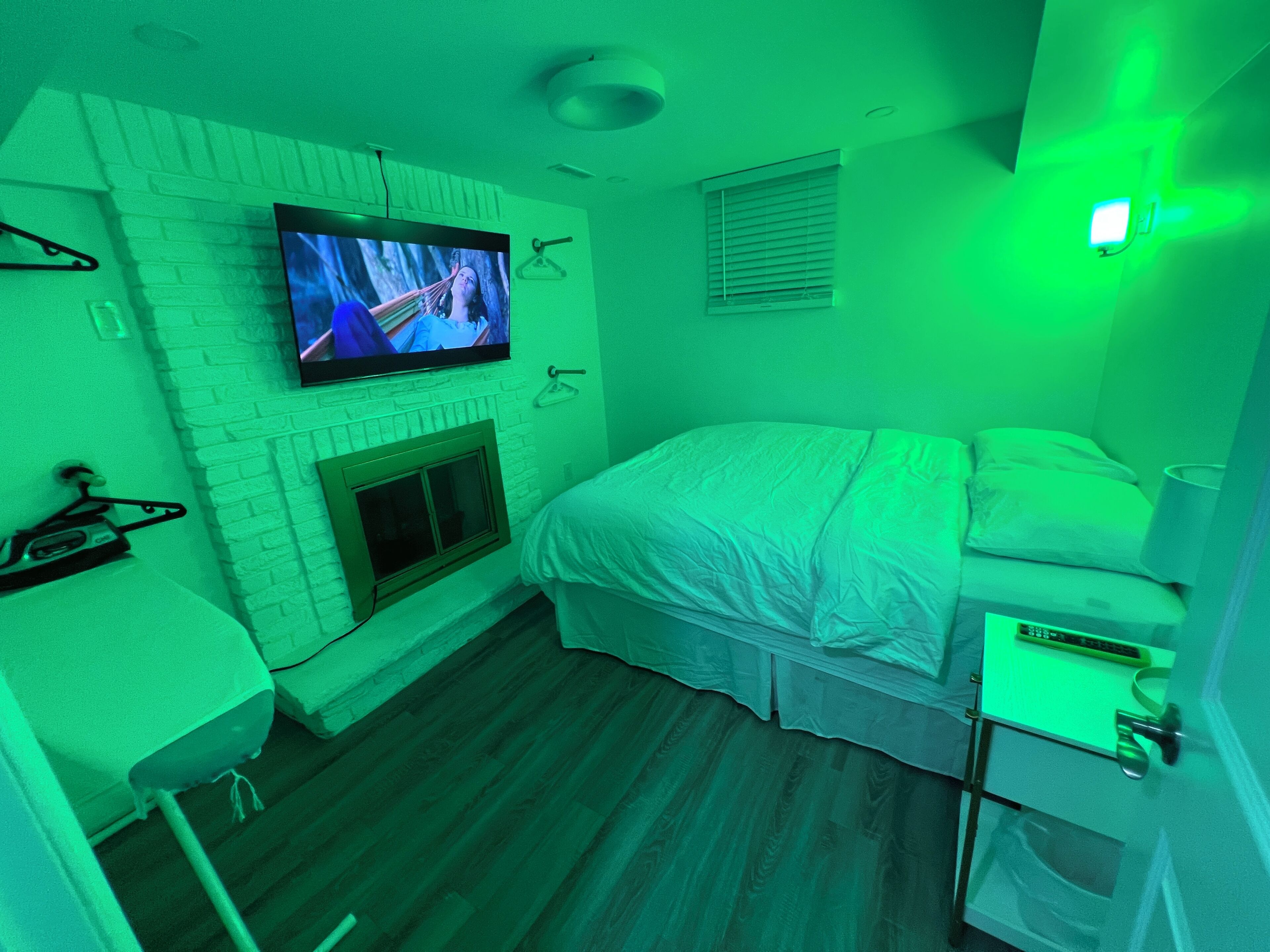 A bedroom with a television mounted on a brick wall, a bed with a white comforter, a bedside table, and a small cabinet.