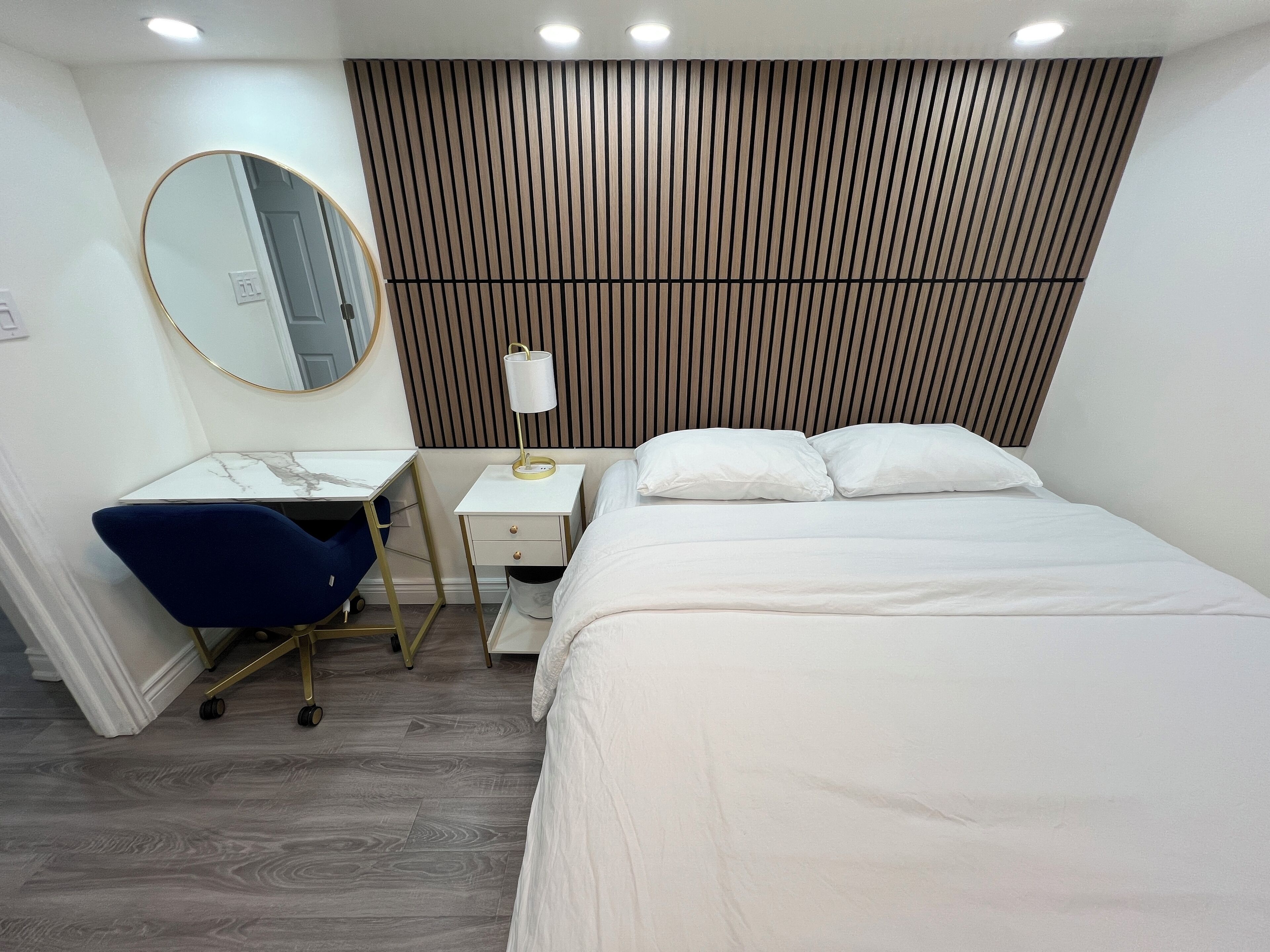A hotel room with a bed, a desk with a chair, a bedside table with a lamp, a mirror, and a wooden panel wall.