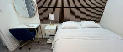 A hotel room with a bed, a desk with a chair, a bedside table with a lamp, a mirror, and a wooden panel wall.