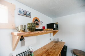 Una cocina con un estante de madera que sostiene una cafetera, una taza y un vaso. Hay un cartel en la pared y un electrodoméstico negro debajo del estante.