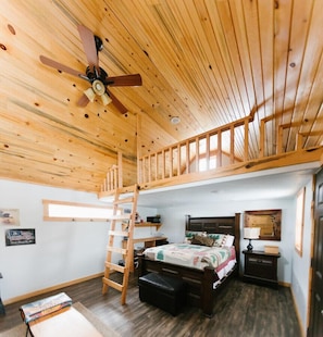 A cabin interior with a loft bedroom, wooden ceiling, a ladder to the upper bunk, a bed with a floral bedspread, a nightstand, and a small desk.