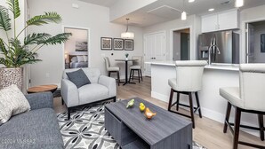 Un salon moderne avec un canapé gris, une table basse et un coin repas comprenant des chaises et une petite table. On y trouve également une cuisine avec des meubles blancs et des appareils électroménagers en acier inoxydable.