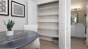 Une pièce avec une table en marbre, une plante en pot et une armoire blanche dont les portes sont ouvertes, laissant apparaître des étagères.