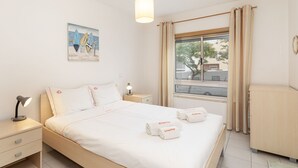 Quarto de hotel bem arrumado, com uma cama grande, uma mesa de cabeceira com abajur, janela com cortinas e um quadro na parede.