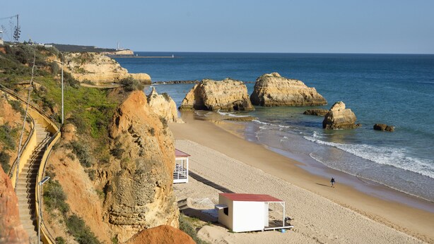 Uma praia costeira com falésias rochosas, um farol ao longe e uma pequena cabana de praia.