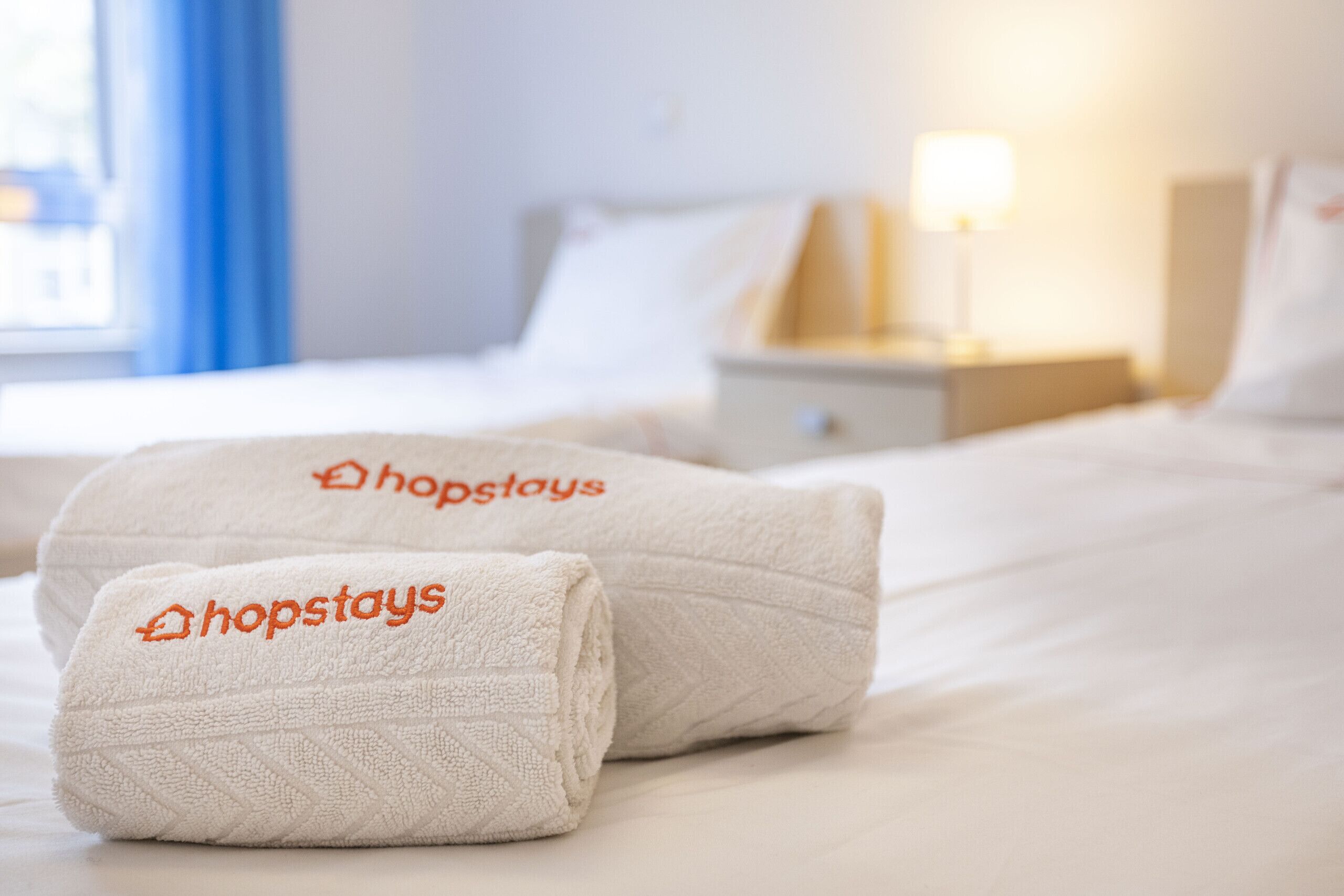 Dos toallas dobladas ordenadamente con el logotipo de 'hopstays' sobre una cama en una habitación de hotel.