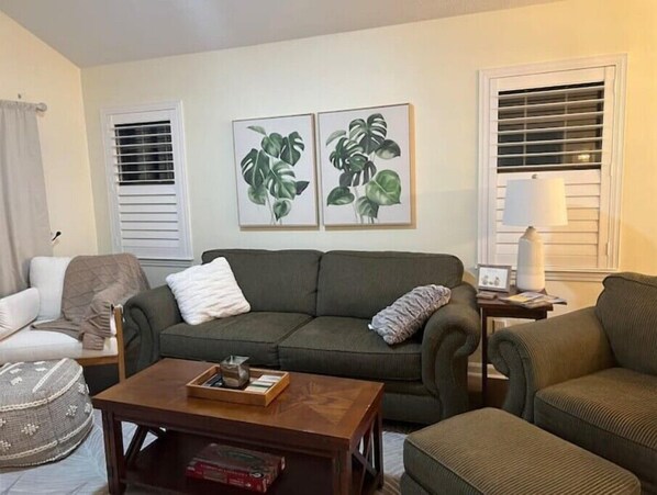 A living room with a green sofa, a wooden coffee table, and a white armchair. There are two framed plant prints on the wall. - Sunsets & Sand Traps on the 17th
(Pawleys Island)