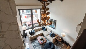 A modern living room with a stone wall, a large window, a wooden ceiling, a beige sectional sofa, a coffee table, and a unique chandelier.