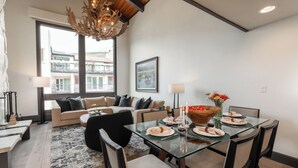 A modern dining area with a glass table, black chairs, and a chandelier. There is a sofa set in the background and a large window with a view of buildings.