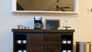 A dark wooden cabinet with multiple shelves holding white mugs, a coffee maker, a framed quote, and a decorative lamp. - Gem of Jacksonville, near NC Beaches & Military Base (Jacksonville)