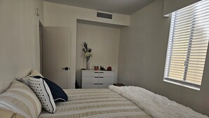 A bedroom with a bed, a dresser, a window with blinds, and a door leading to another room.