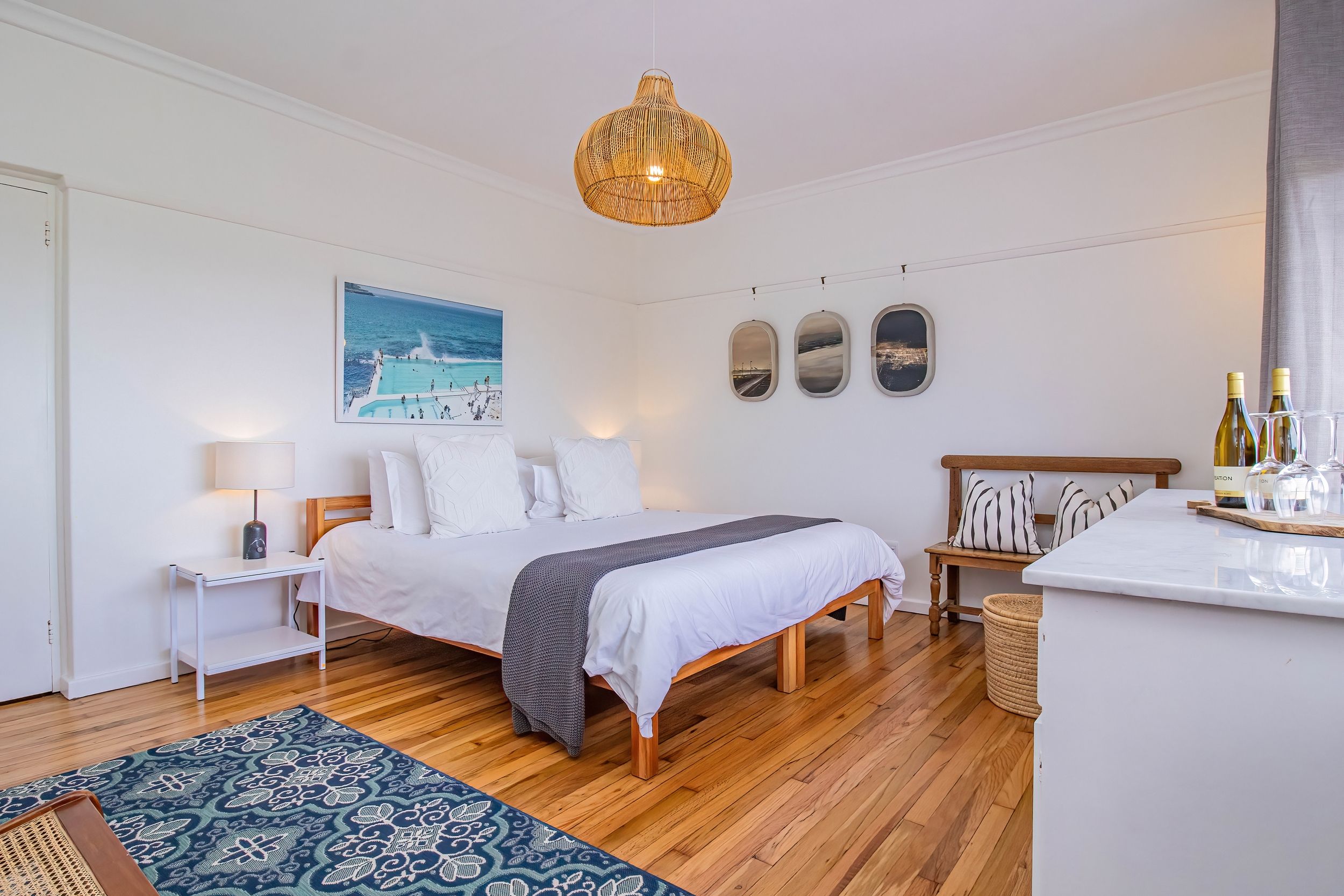 A hotel room with a bed, bedside tables, a desk, a woven chair, a woven basket, a hanging light fixture, and a painting of a beach scene.