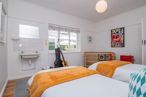 A room with two beds, a window, a guitar, a sink, and a colorful artwork on the wall. - Fairview Hermanus | Ocean View Home, Sleeps 8 (Hermanus)