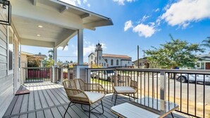Une terrasse en bois, agrémentée de meubles en osier et d’une table en verre, qui offre une vue sur une rue bordée de voitures garées et sur une église en arrière-plan.