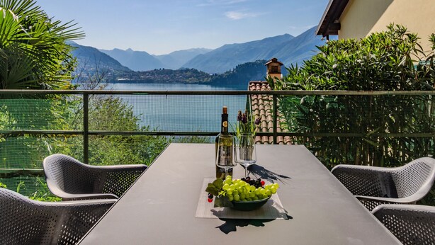 Ein Tisch mit einer Weinflasche, einem Glas und einer Schüssel Trauben auf einem Balkon mit Blick auf einen See und Berge im Hintergrund.
