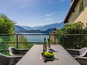 Una mesa con una botella de vino, una copa y un bol de uvas en un balcón con vistas a un lago y montañas al fondo.