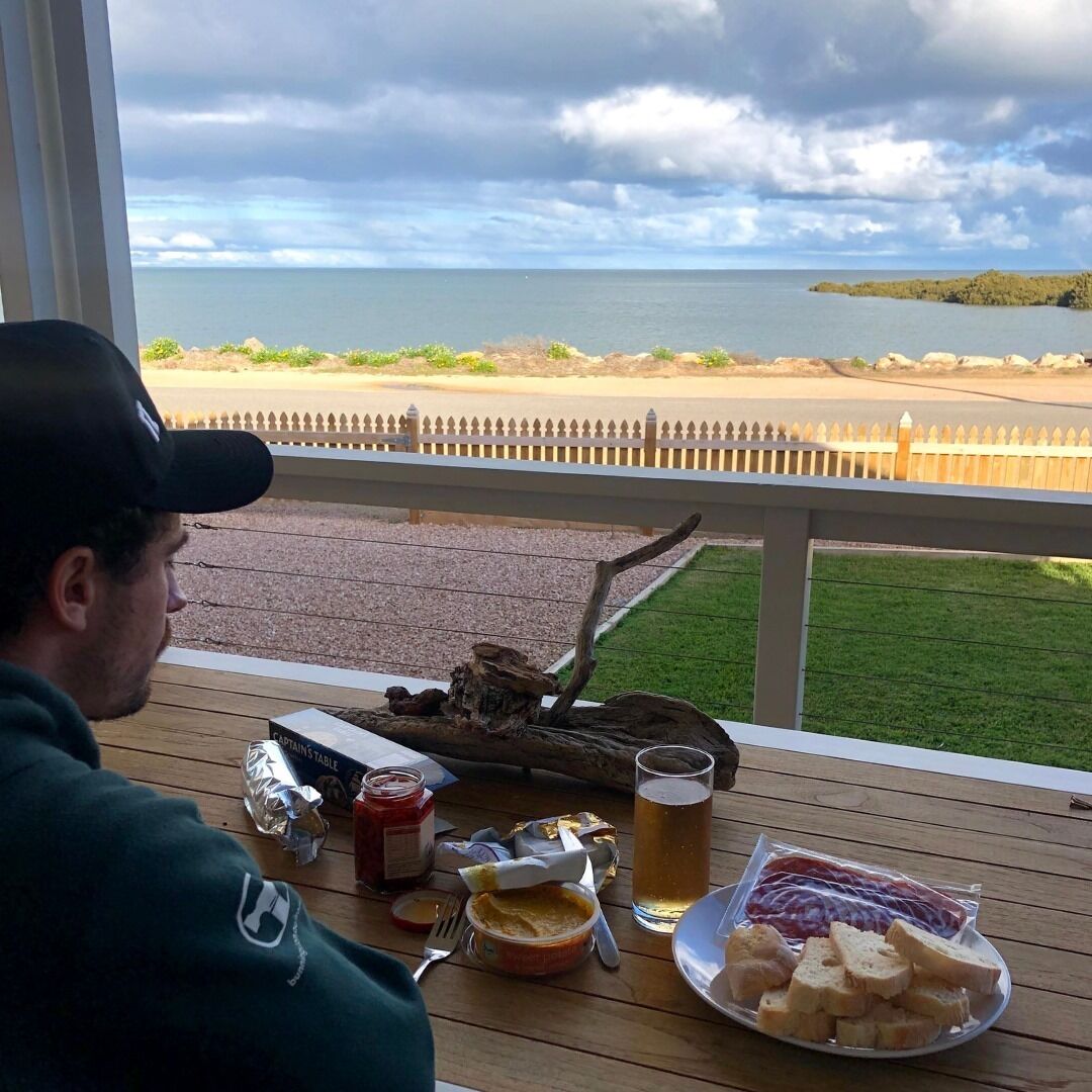 Una mesa de comedor con comida y bebidas, una terraza de madera y vistas al mar y una isla distante.