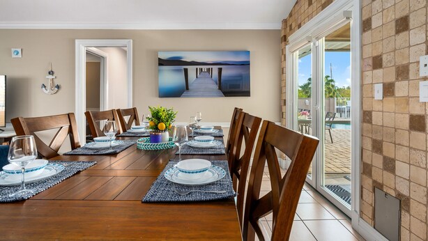 A dining area with a wooden table set for four, a painting of a pier, and a view of a pool through glass doors.