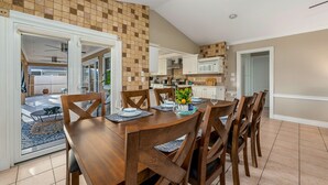 A dining area with a wooden table and chairs, a kitchen in the background, and a view of the outside patio.