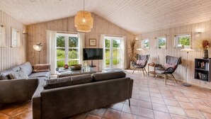 A spacious living room with a large sofa, a coffee table, and a TV. There are two chairs and a bookshelf. The room has wooden walls and a tiled floor.