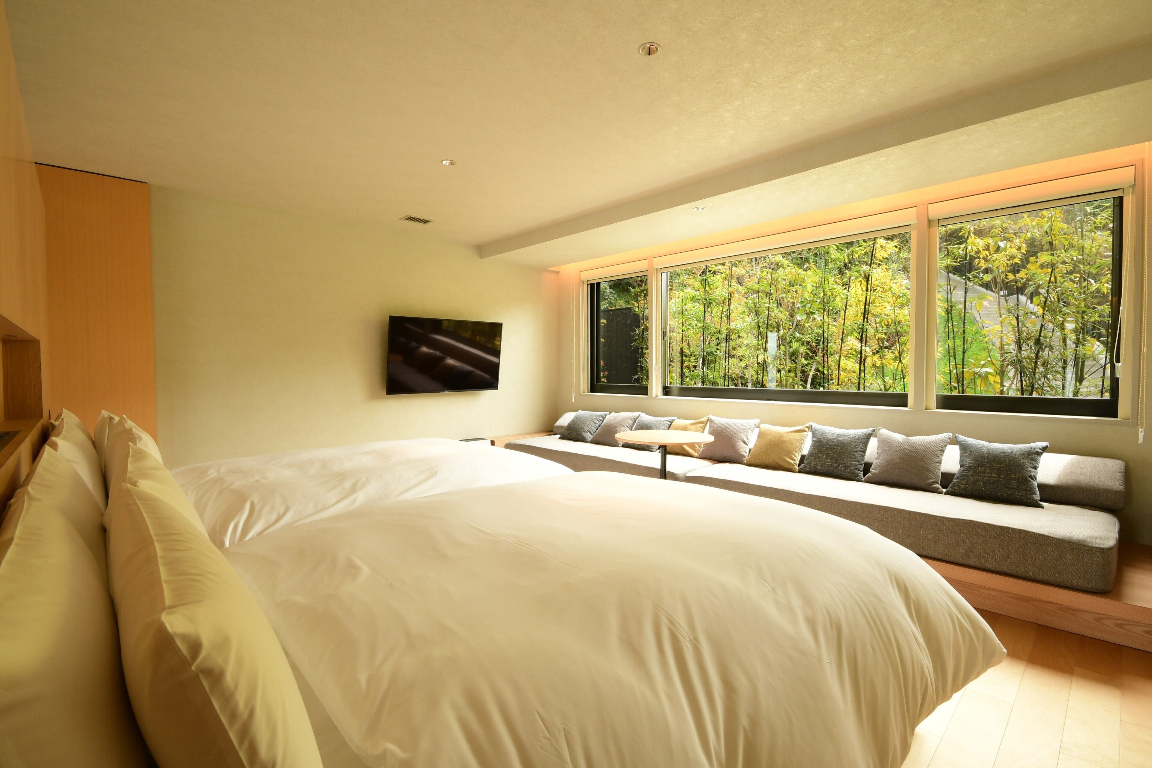 A bedroom with a large bed, a built-in bench with cushions, a TV mounted on the wall, and a window with a view of greenery.