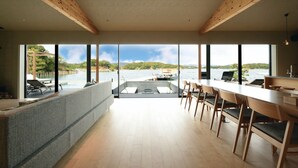 A modern interior with large windows, a long dining table, and a seating area with a view of a lake and boats. - MOKU ISESHIMA (Shima)