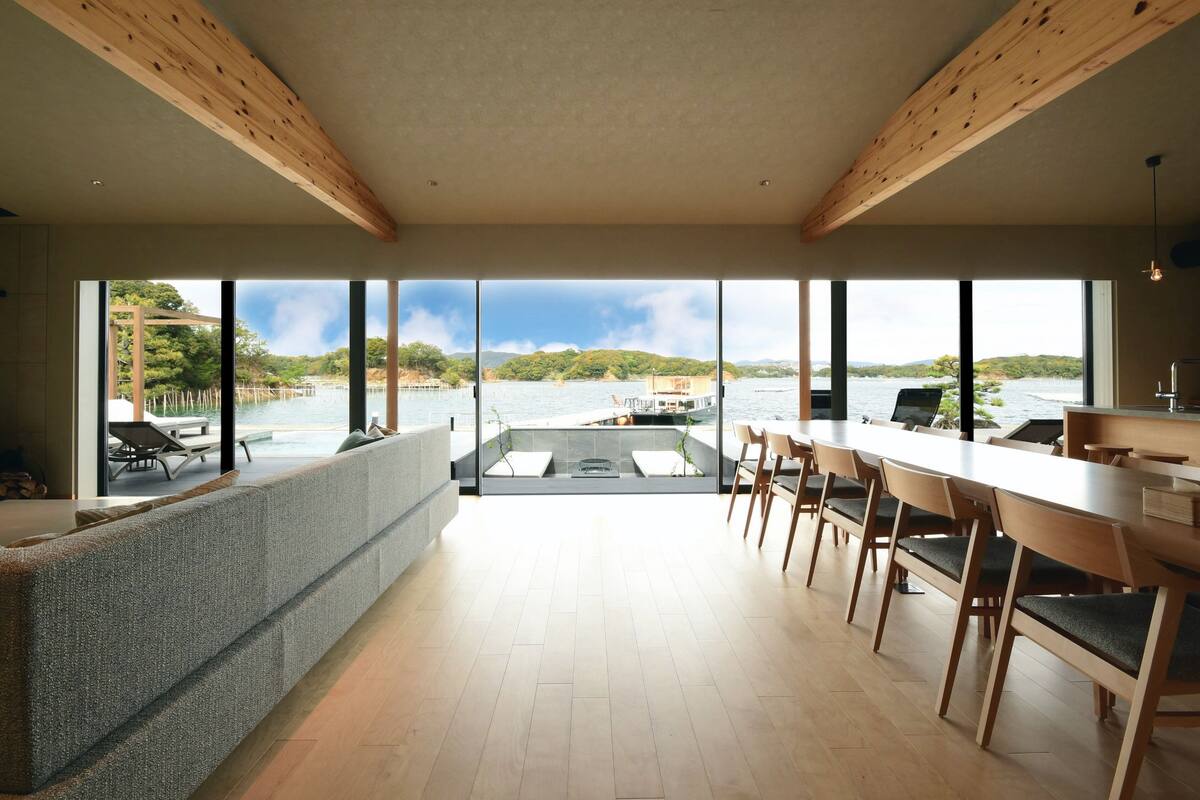 Un interior moderno con ventanas amplias, una larga mesa de comedor y una zona de estar con vista a un lago y barcos.