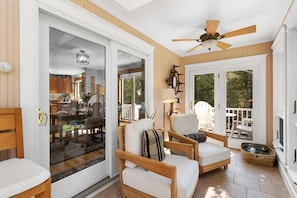 Una stanza luminosa con ventilatore a soffitto, zona pranzo con tavolo e sedie, e vista all'esterno attraverso una porta in vetro.
