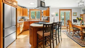Une cuisine dotée de meubles en bois, d’un réfrigérateur en acier inoxydable, d’un îlot central avec des chaises et d’un coin repas avec une table et des chaises.