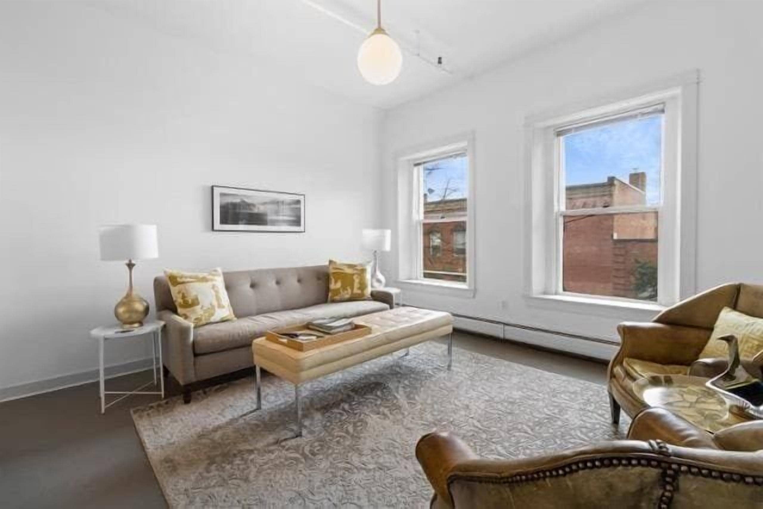 Een woonkamer met een bank, fauteuil, salontafel en vloerkleed. Er zijn twee ramen en een schilderij aan de muur.
