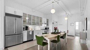 A modern kitchen with stainless steel appliances, a dining area with a wooden table and green chairs, and a visible clock on the wall. - Rockland Central Over Main, downtown living (Rockland)