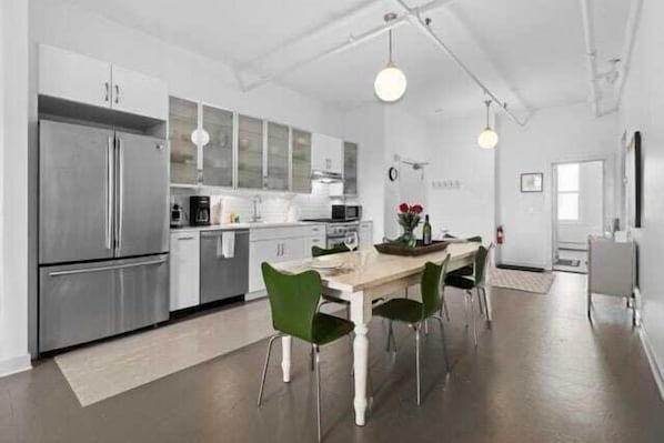 A modern kitchen with stainless steel appliances, a dining area with a wooden table and green chairs, and a visible clock on the wall.