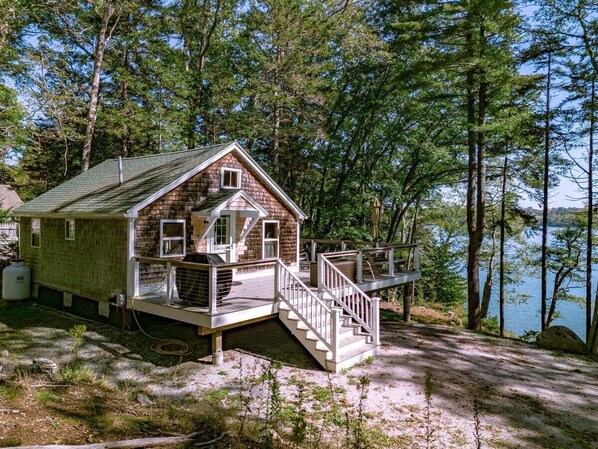 A cabin with a deck, a staircase, and a view of a lake surrounded by trees. - Settlers Cottage, cute Maine home with water views (Cushing)