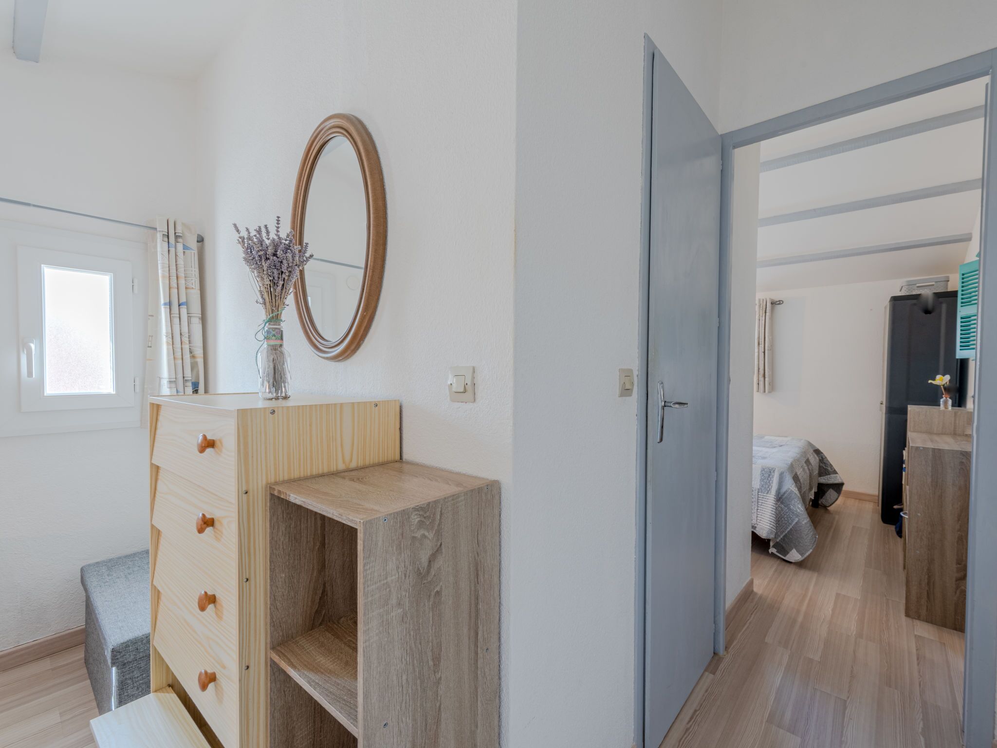 A room with a wooden dresser, a mirror, and a window. There is a door leading to another room with a bed and a wardrobe.