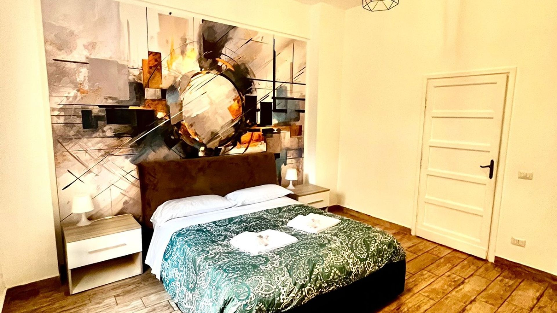 Une chambre à coucher avec un lit, une table de chevet, une lampe, une porte et un mur orné d’œuvres d’art abstraites.