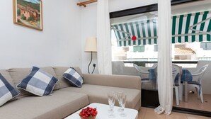 Una sala de estar con un sofá beige, dos almohadas a cuadros, una mesita blanca con un frutero y vasos, y vistas a un balcón con un toldo a rayas y mobiliario exterior.