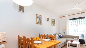 Un comedor con una mesa de madera, sillas y un frutero. Hay un sofá y una pequeña mesa blanca con vasos y un cuenco de fruta.