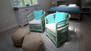 Quarto com teto inclinado, uma janela grande, cama, duas cadeiras de vime, cômoda e mesa de cabeceira.